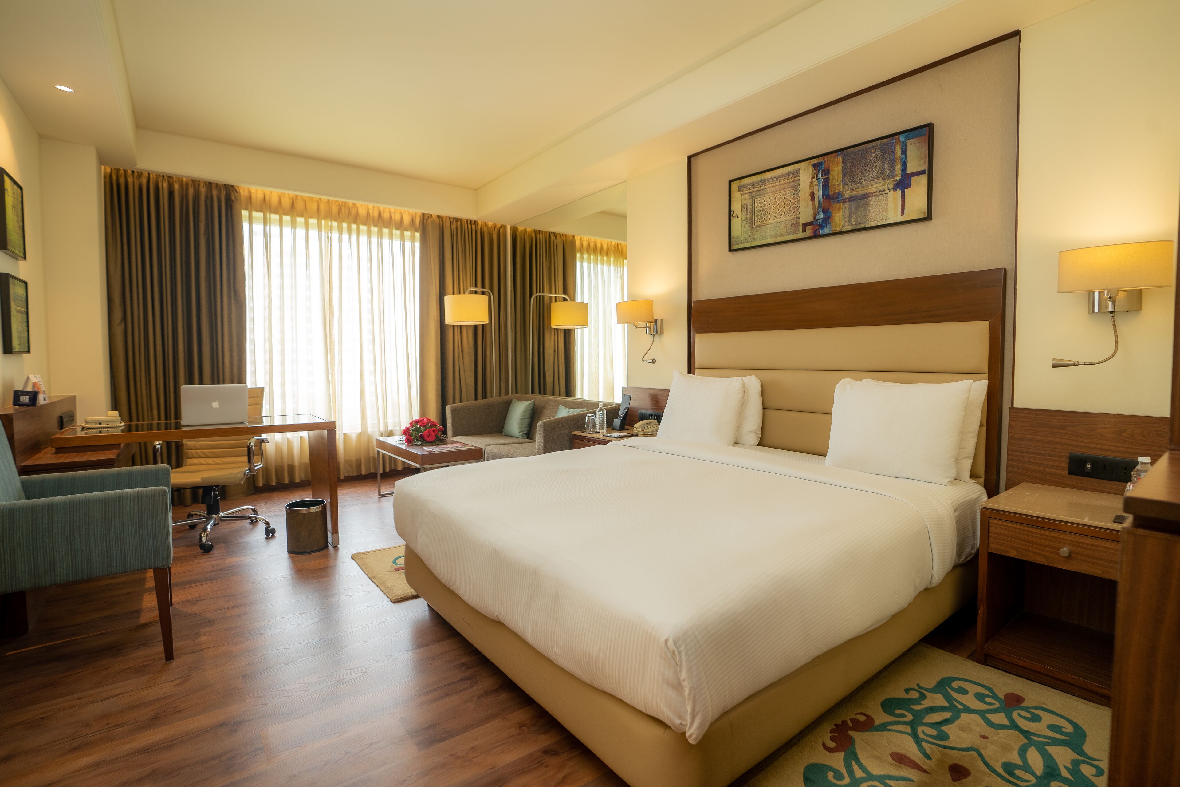 A hotel room with a large bed, a desk with a laptop, a chair, and a small table with a lamp.