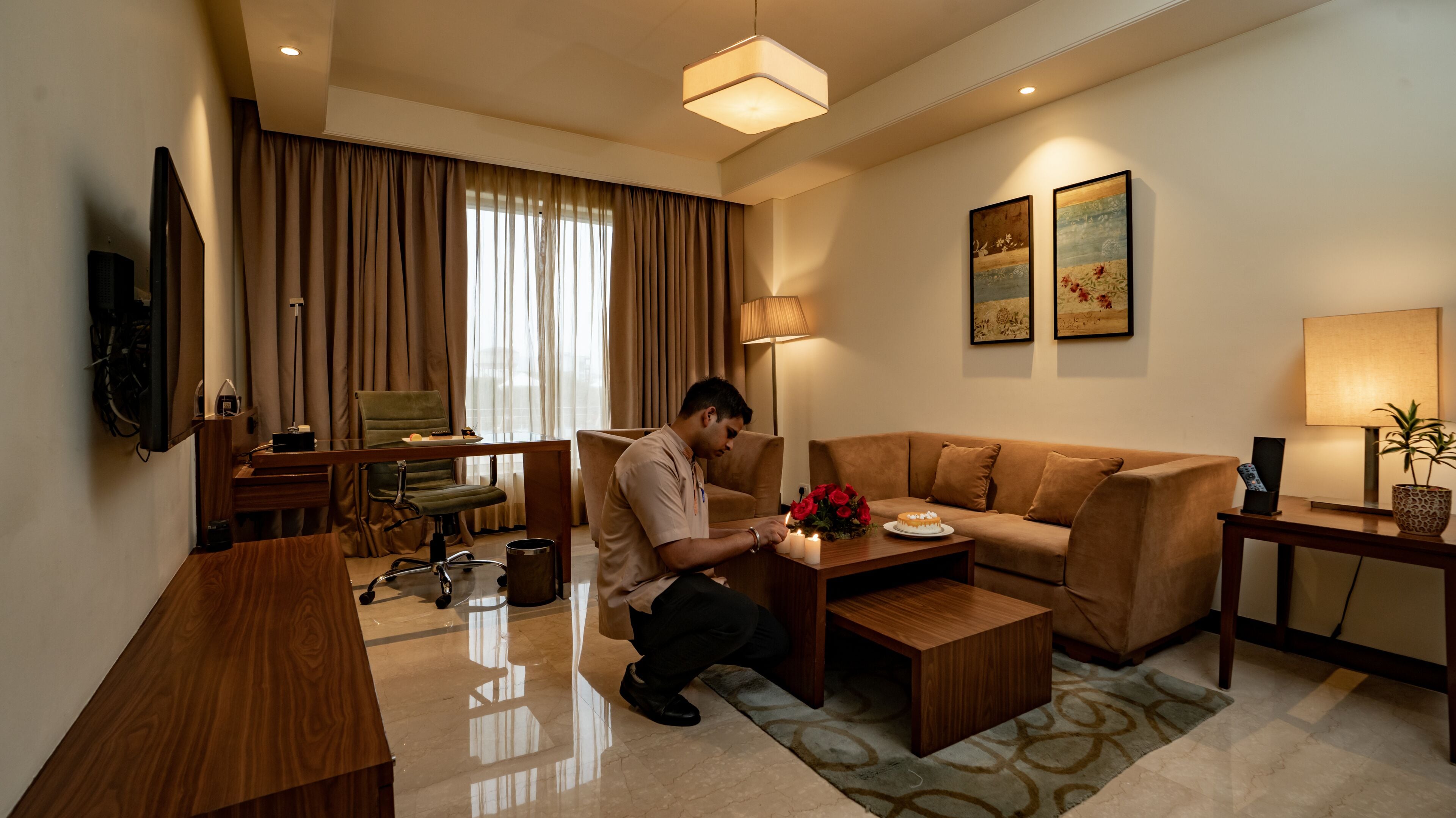 A modern hotel room with a sofa, coffee table, and desk. There is a television mounted on the wall, a lamp, and a potted plant.