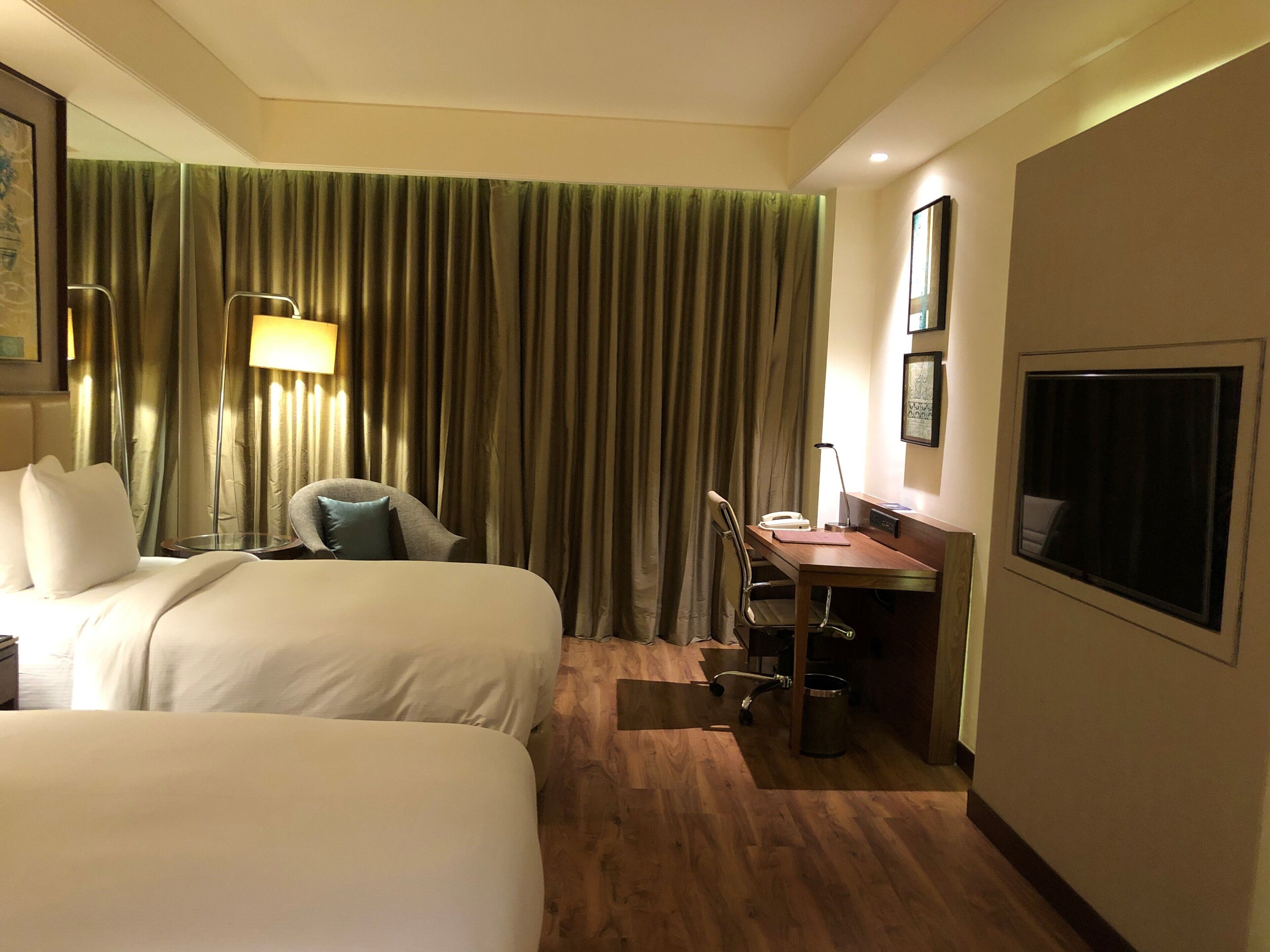 Una camera d'albergo con due letti, una scrivania, una sedia, una lampada e un'ampia finestra.