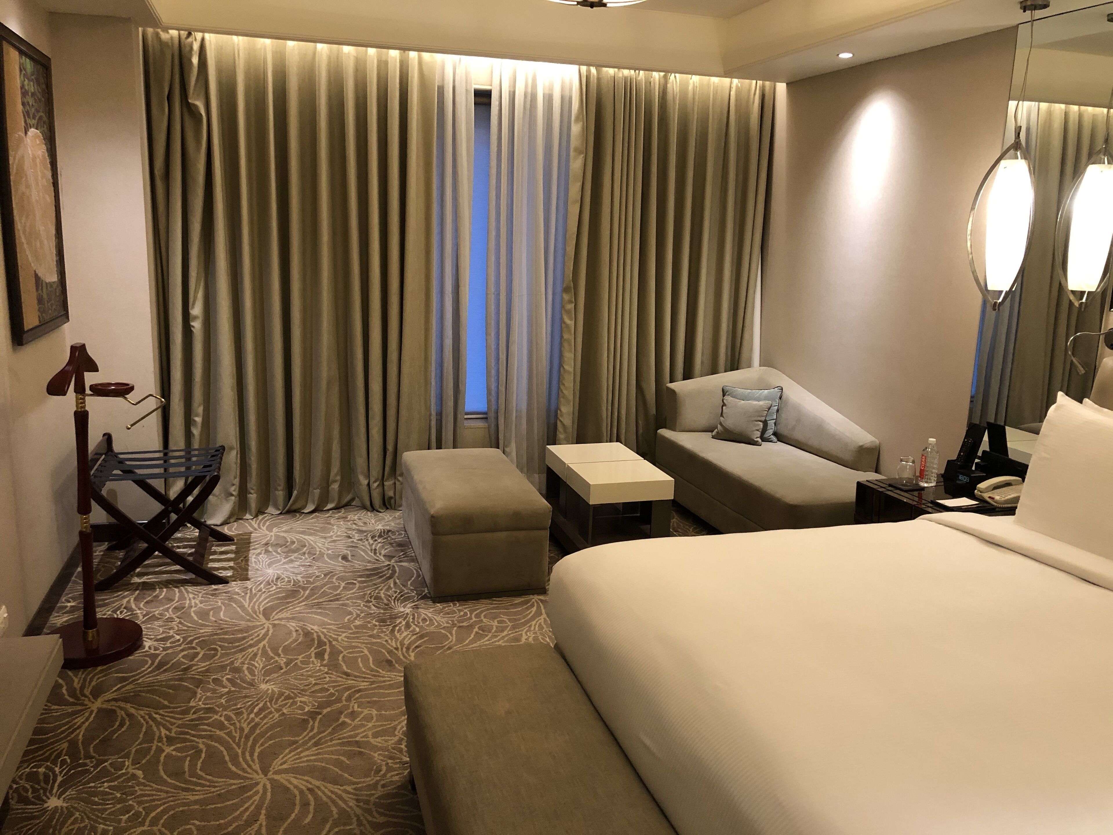 A hotel room with a large bed, a sofa, a small table, a bench, and a chair. There is a window with curtains, a mirror, and a lamp.