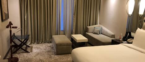 A hotel room with a large bed, a sofa, a small table, a bench, and a chair. There is a window with curtains, a mirror, and a lamp.