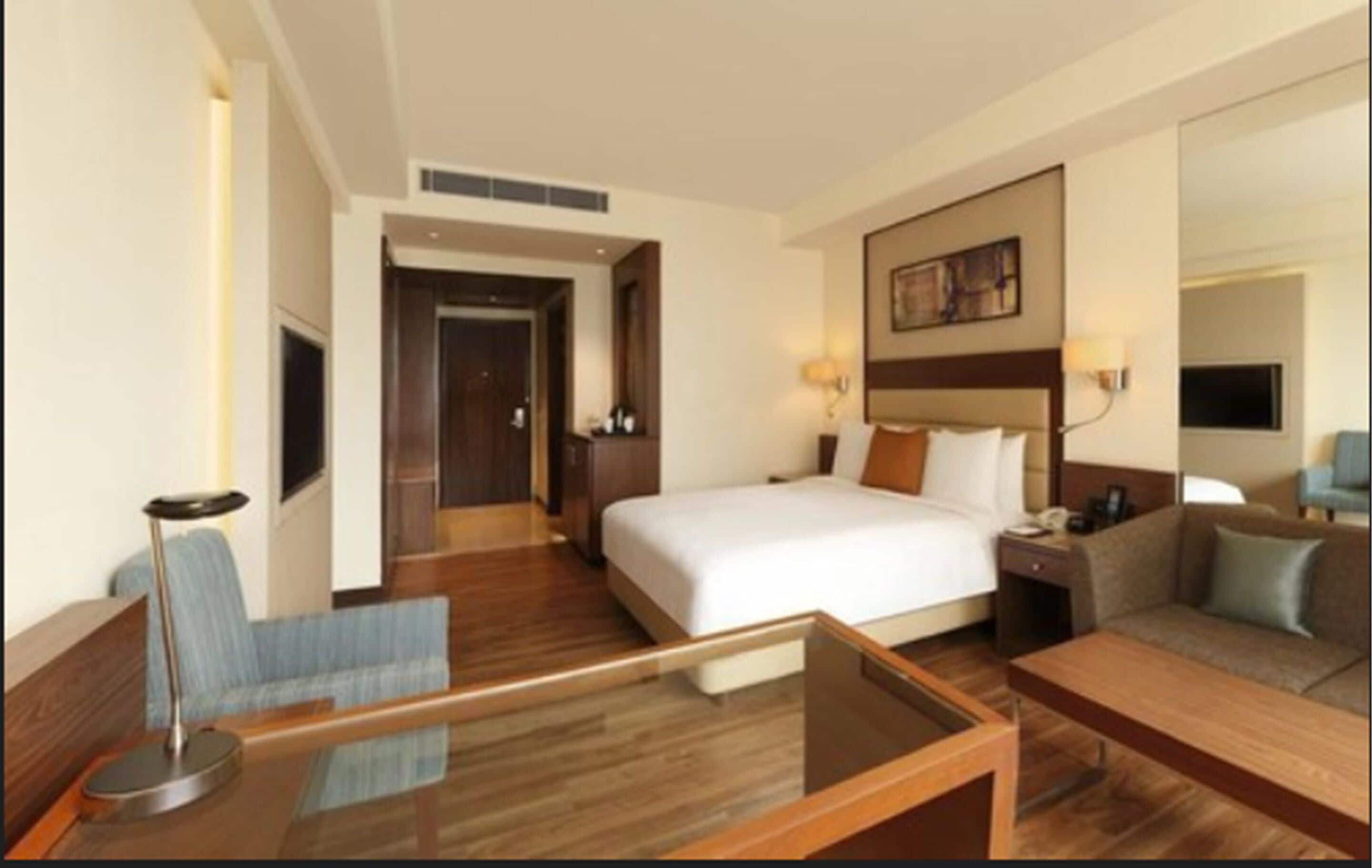 Quarto de hotel com uma cama grande, um sofá, uma escrivaninha com cadeira e uma mesa de centro de vidro.