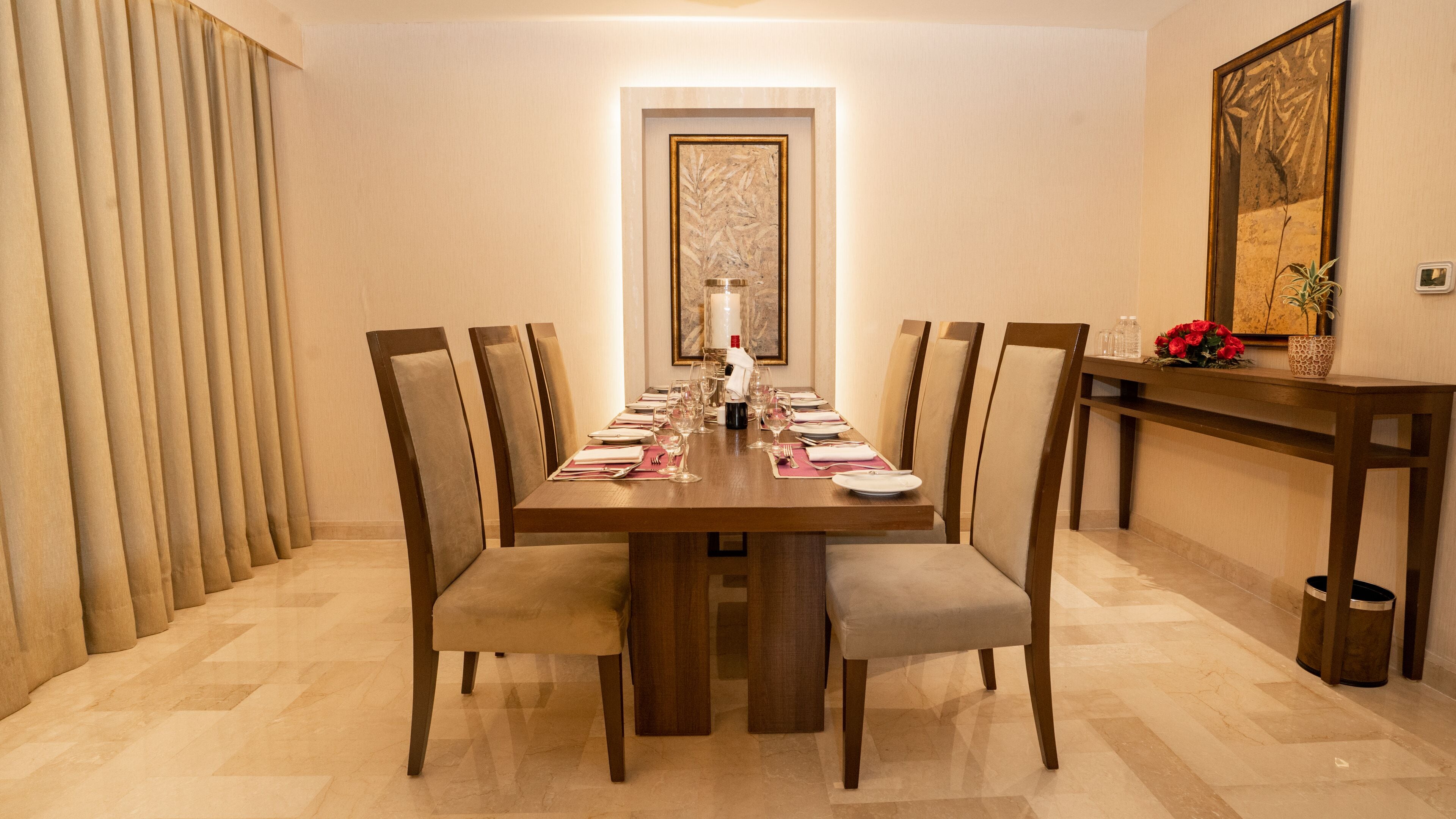 Comedor con una larga mesa dispuesta para seis comensales, sillas a los lados, un jarrón con flores y una obra de arte enmarcada en la pared.