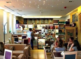 Um café acolhedor com móveis de madeira, um balcão com cardápio e prateleiras repletas de lanches e bebidas.