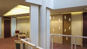 Moderni hotellin aula, jossa on korkea katto, upotetut valaisimet sekä oleskelualue, jossa on tuoleja ja pieni pöytä.