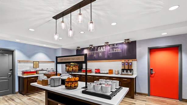 A modern breakfast area with a white counter, hanging pendant lights, and a red door. - Hampton Inn Omaha/West Dodge Road (Old Mill) (Omaha)