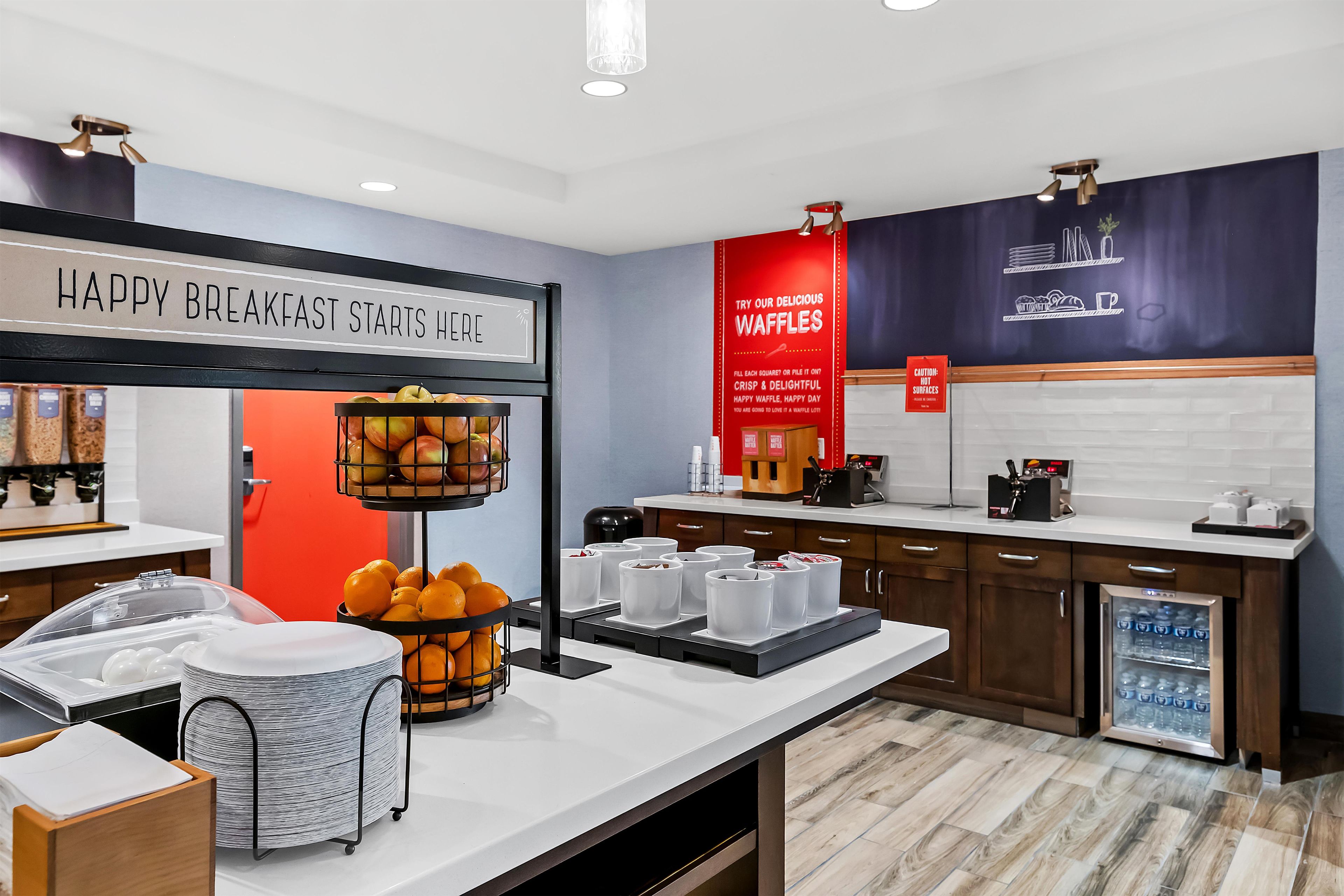 A breakfast area with a sign that reads 'HAPPY BREAKFAST STARTS HERE,' a fruit display, a waffle station, and a coffee machine.