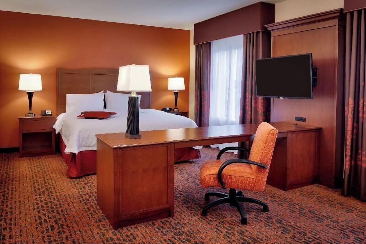 A hotel room with a bed, two bedside tables with lamps, a desk with an orange chair, a flat-screen TV mounted on the wall, and a patterned carpet.