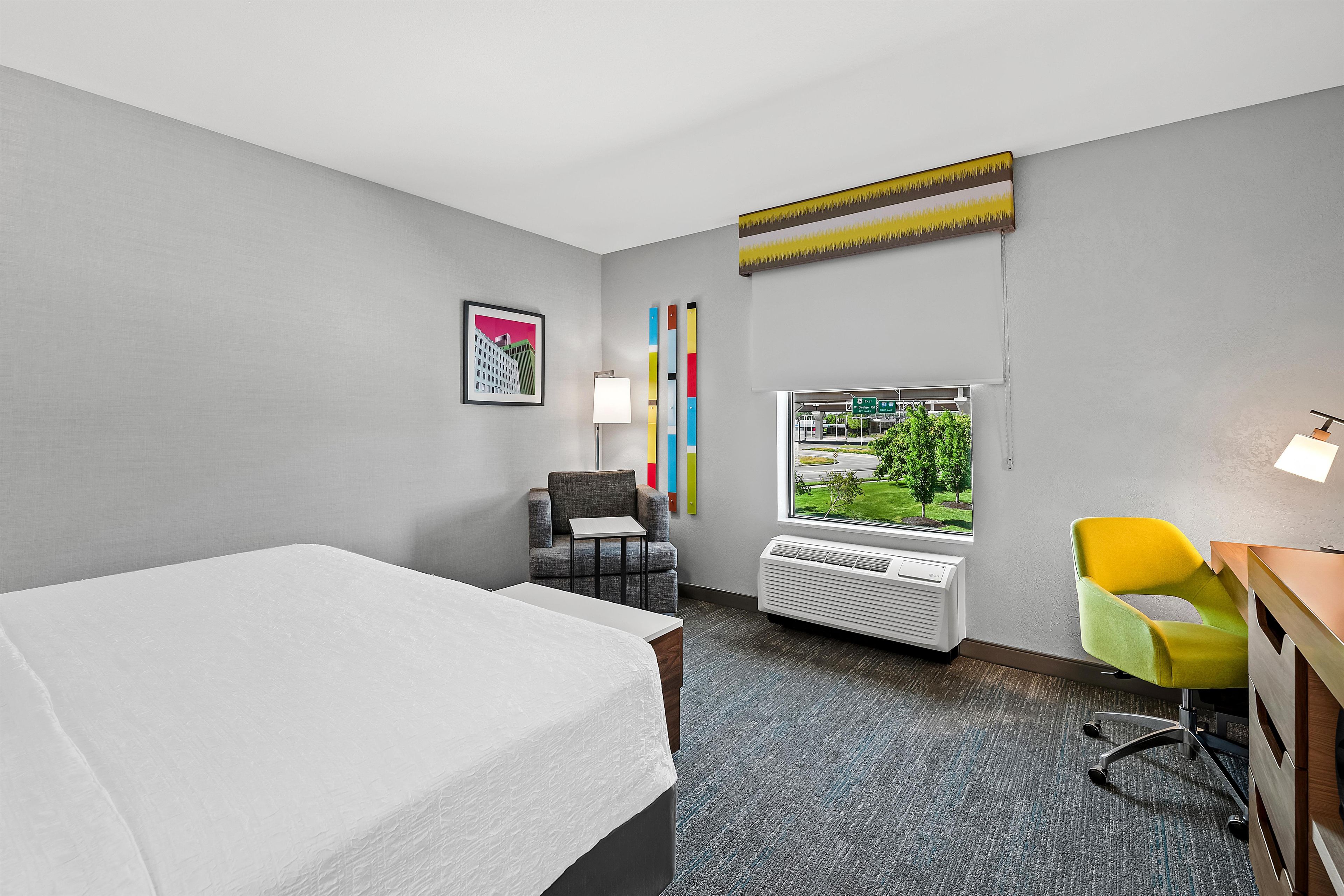 A hotel room with a bed, a desk, a chair, a window with a view, a lamp, and a wall-mounted air conditioner.