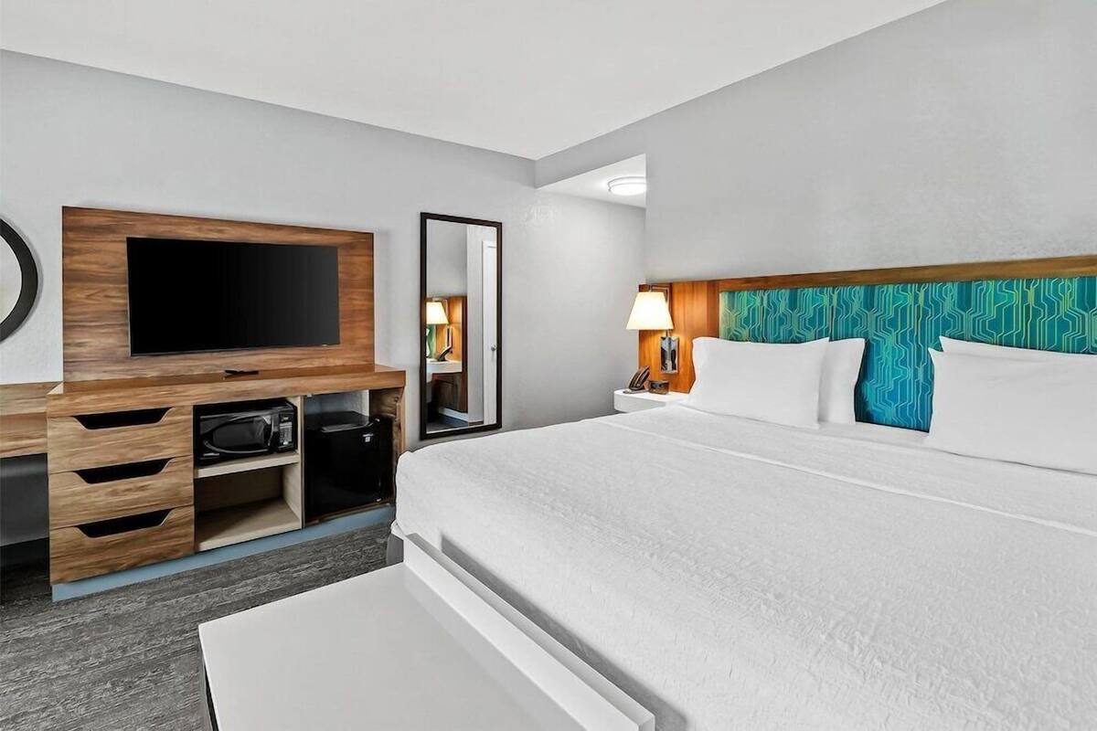 A hotel room with a large bed, a TV mounted on a wooden cabinet, a bedside table with a lamp, and a mirror.