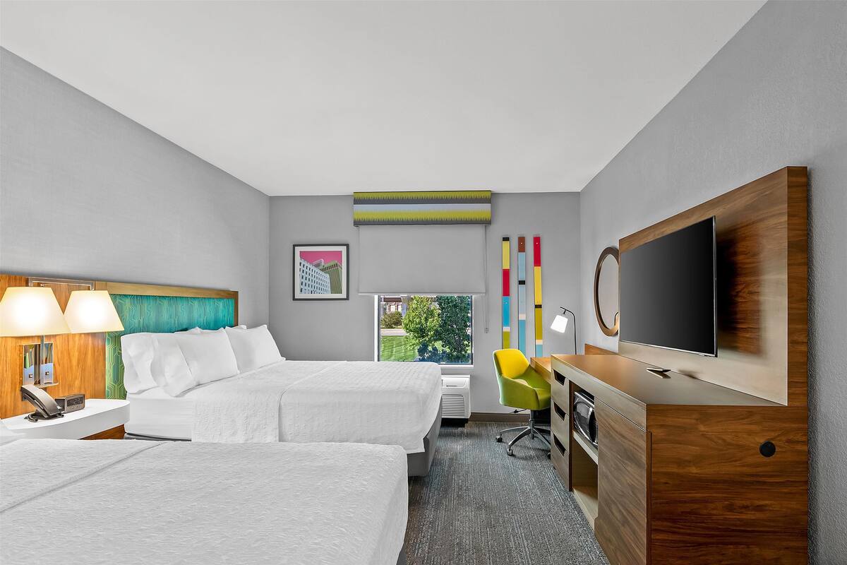A hotel room with two beds, a desk, a TV, and a window with a view of greenery.