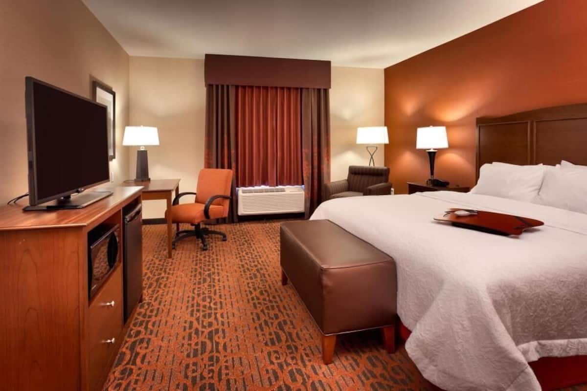A hotel room with a bed, a desk with a TV, a chair, a lamp, and an air conditioning unit.