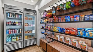 A snack and beverage vending area with refrigerated and frozen sections, shelves stocked with various snacks and drinks, and a wooden counter with additional snack options. - Hampton Inn Omaha/West Dodge Road (Old Mill) (Omaha)