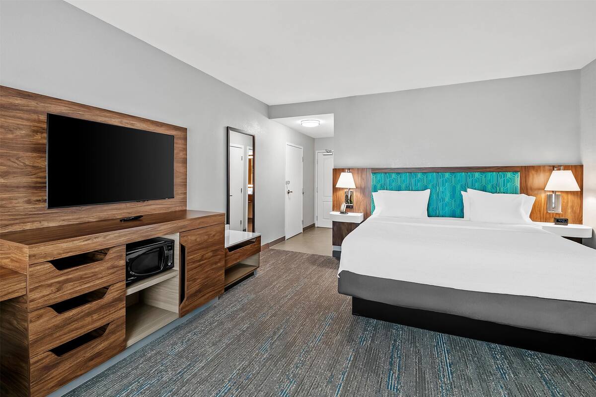A modern hotel room with a large bed, a flat-screen TV, a wooden TV stand, a microwave, a nightstand with lamps, and a bathroom door.