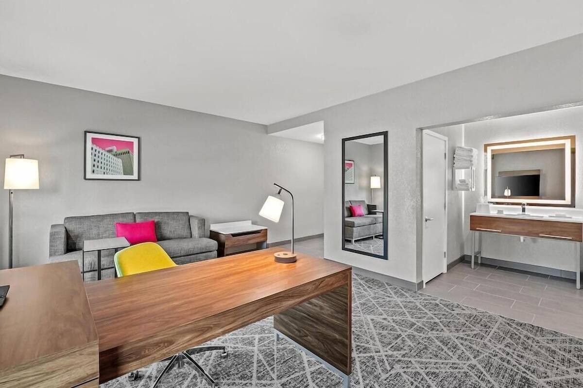 A modern hotel room with a wooden desk, a grey sofa, a small bedside table, a lamp, a mirror, and a bathroom with a vanity and a shower.