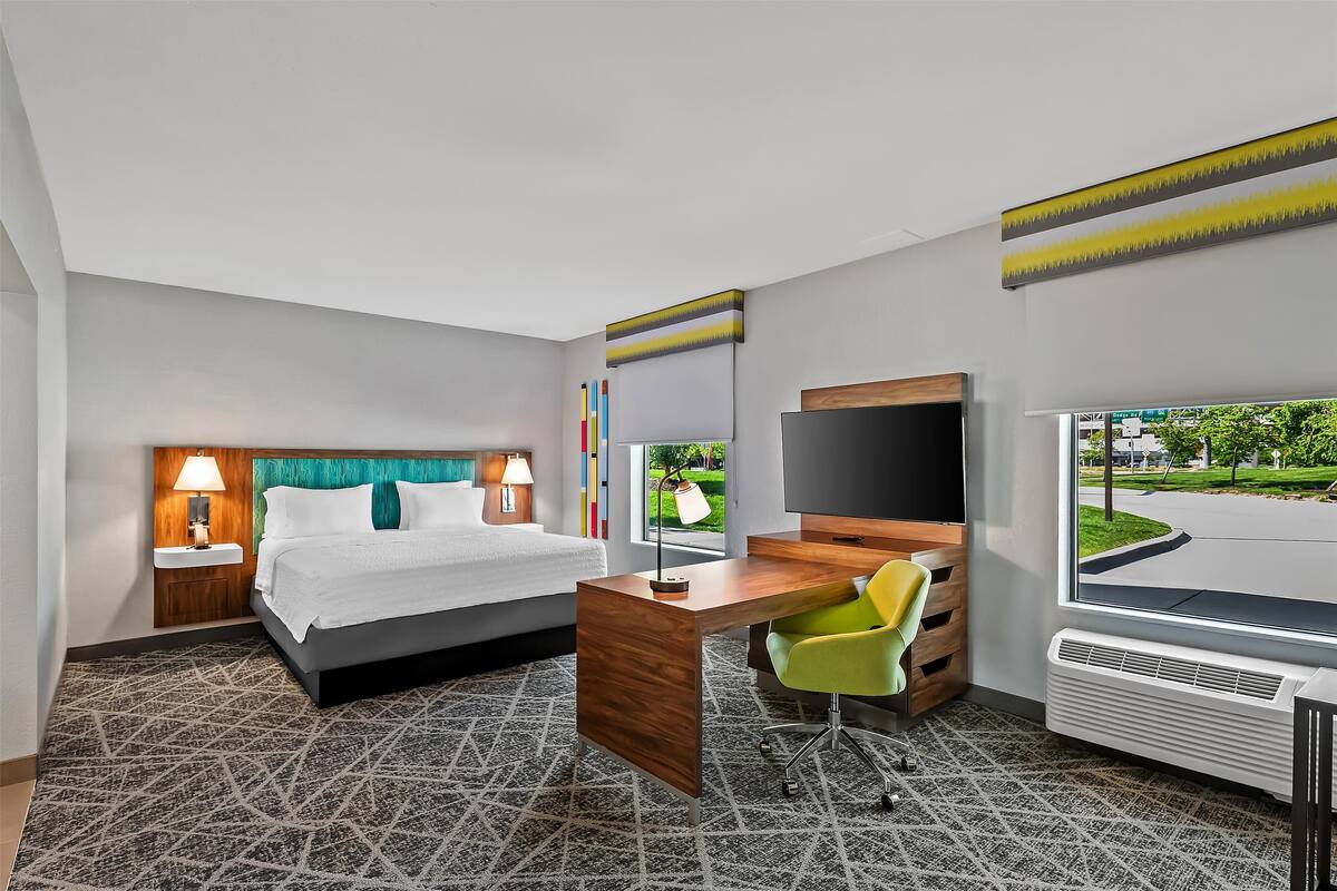 A hotel room with a bed, desk, chair, TV, and a window with a view of a green landscape.