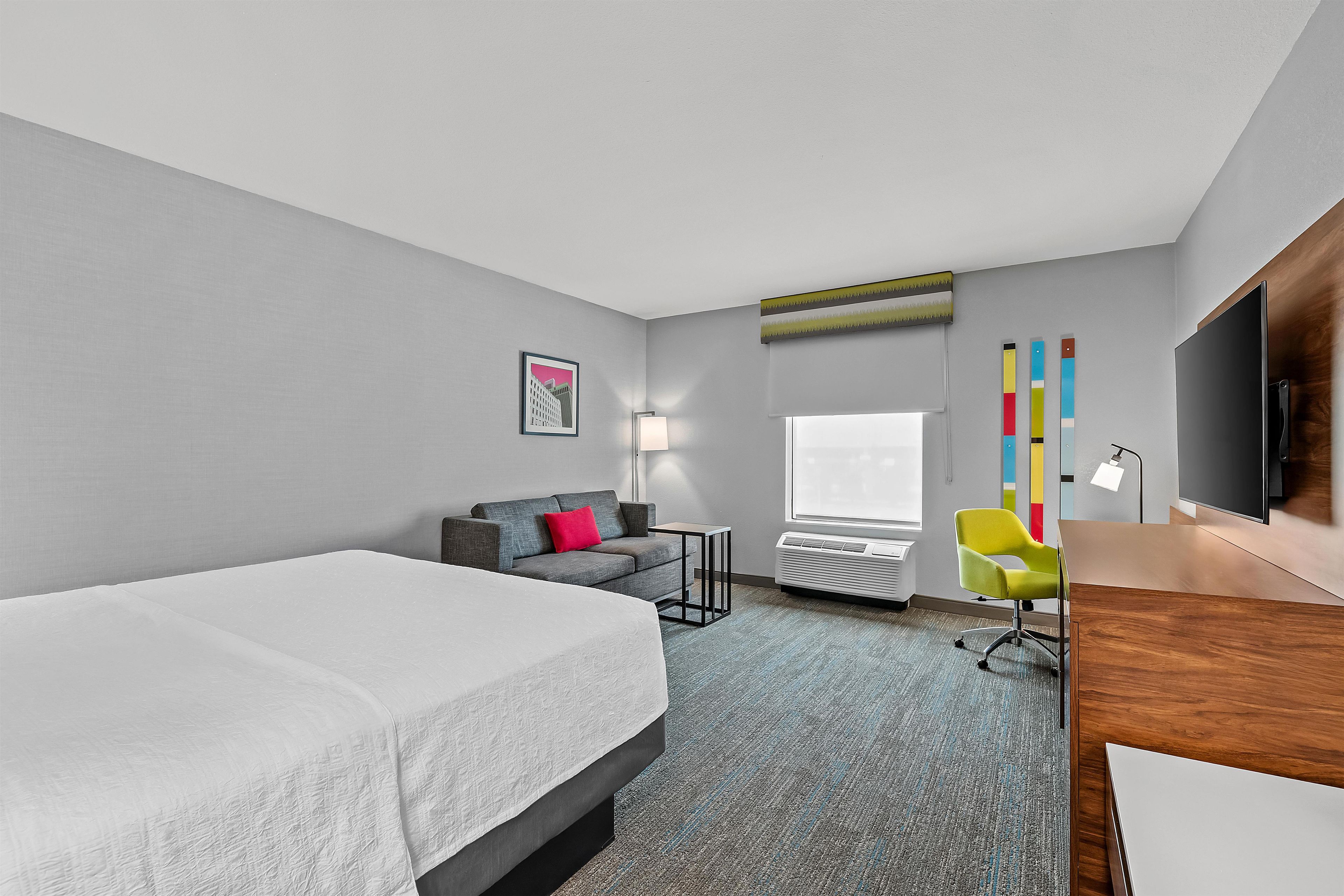 A hotel room with a bed, a gray sofa, a desk, a chair, a TV, and a window.