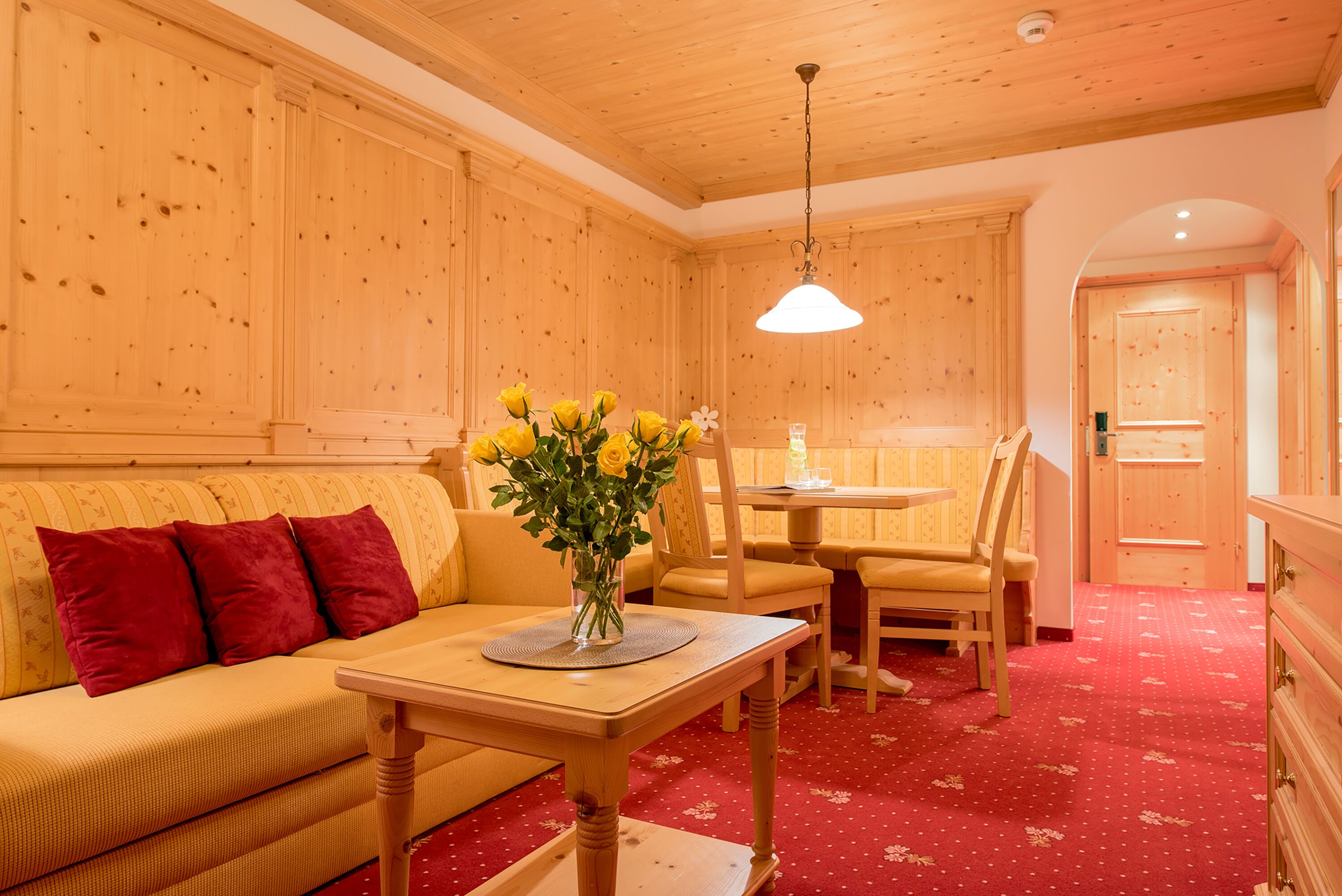 Una habitación acogedora con paredes de madera y alfombra roja, que incluye un sofá, una mesa de comedor con sillas y un jarrón con flores amarillas.