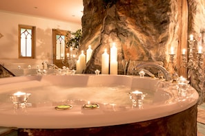 A large, oval-shaped bathtub with multiple candles and glass containers, set against a backdrop of a stone wall and a mirror. - Hotel Bergkristall (Silbertal)