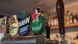 A bar tap with various beer brands, a metal pipe, and a blurred background of shelves with bottles. - Ski Rider Hotel (Wilsons Valley)