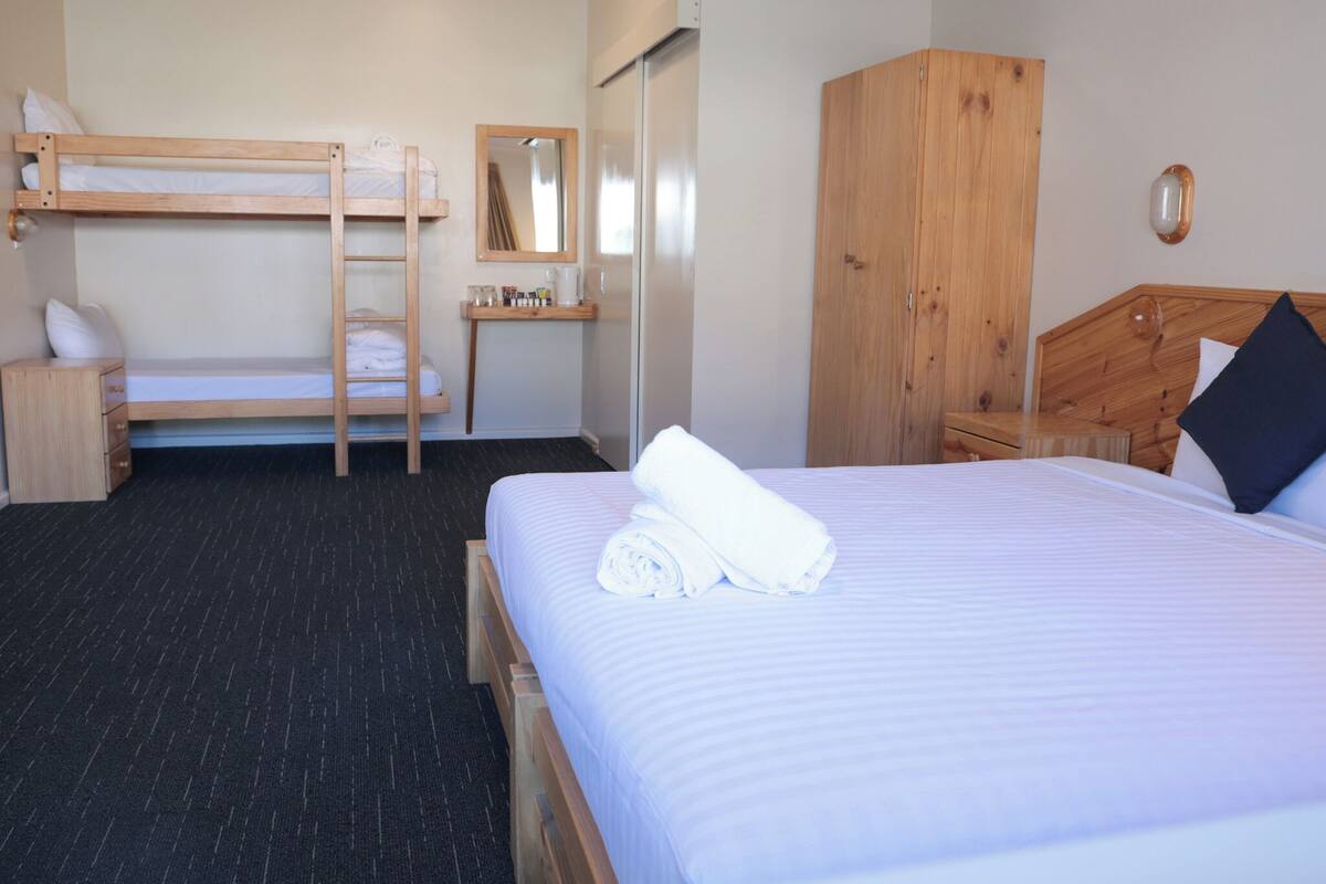 A hotel room with a bunk bed, a wooden wardrobe, a bedside table, a mirror, and a neatly made bed.