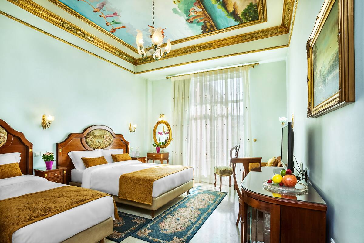 A hotel room with two beds, a desk with a fruit bowl, a mirror, and a chandelier.