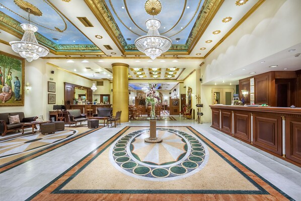 A grand lobby with a central fountain, ornate chandeliers, and a detailed ceiling mural.