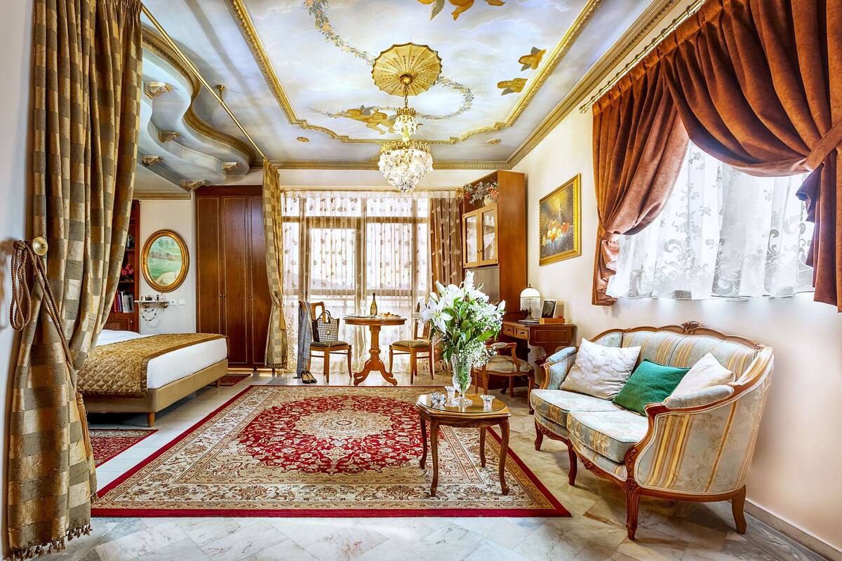 A spacious room with a large bed, a sofa, a chandelier, and a red and white patterned rug.