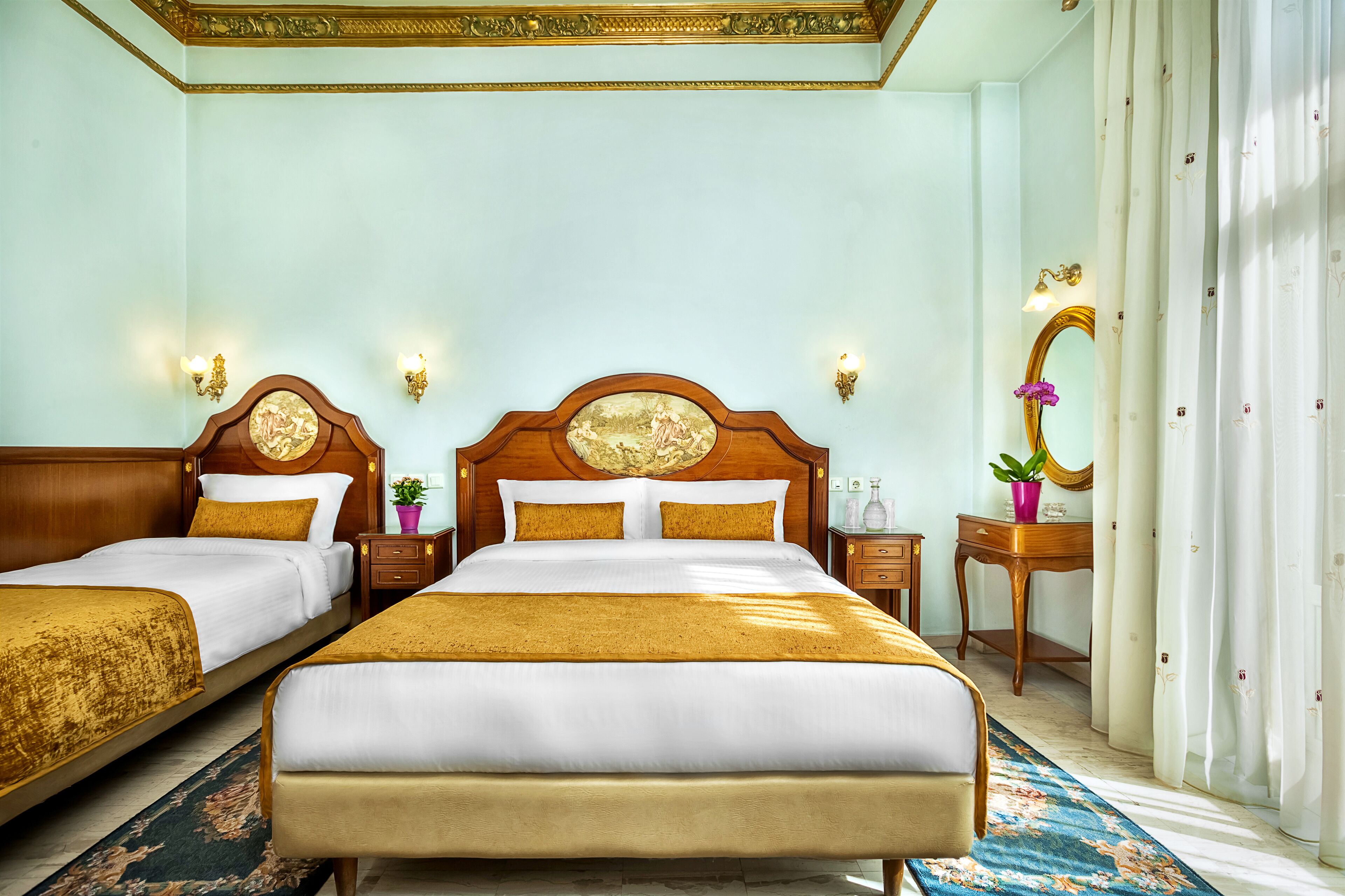 A hotel room with two beds, a wooden headboard, a nightstand with a lamp, a small table with a plant, and a mirror with a floral curtain.