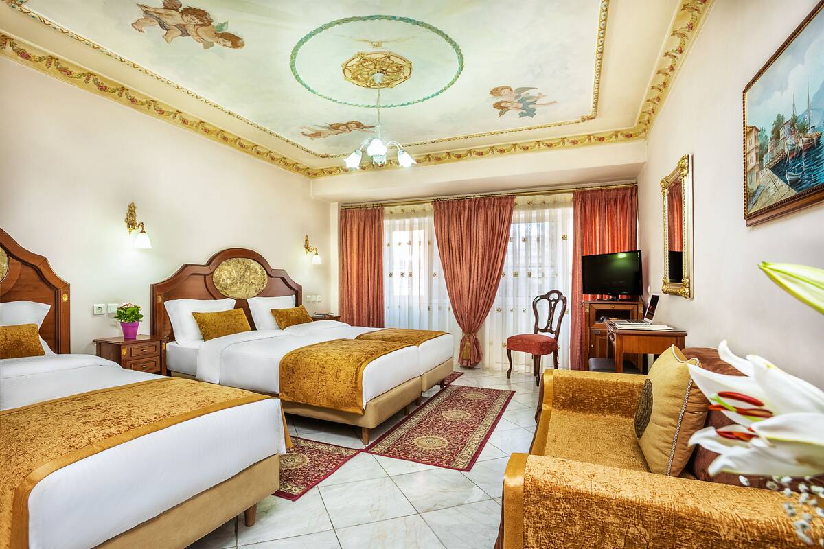 A hotel room with two beds, a desk with a computer, a TV, and a large window with curtains.
