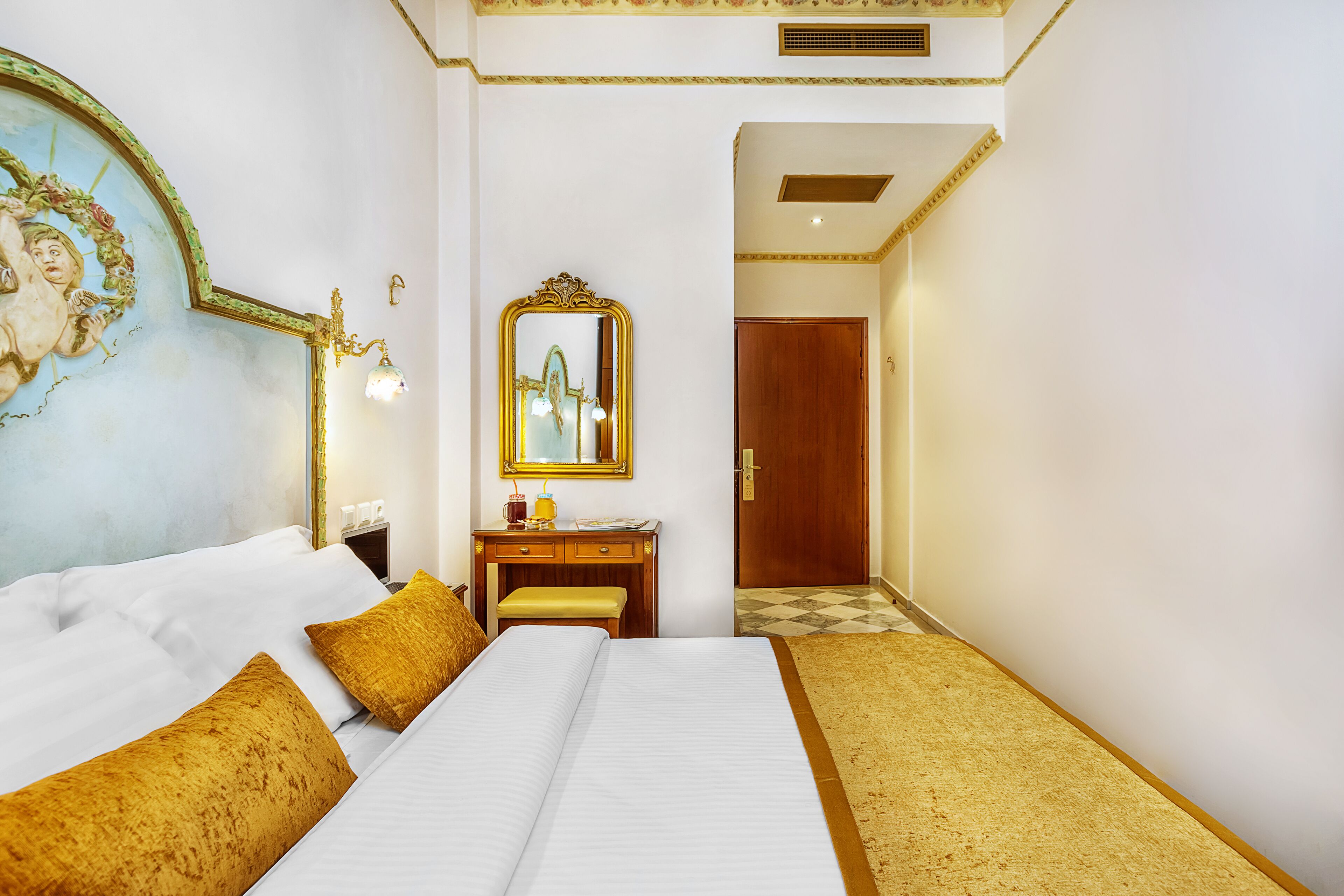 A hotel room with a large bed, a wooden desk, a mirror, and a door leading to another room.