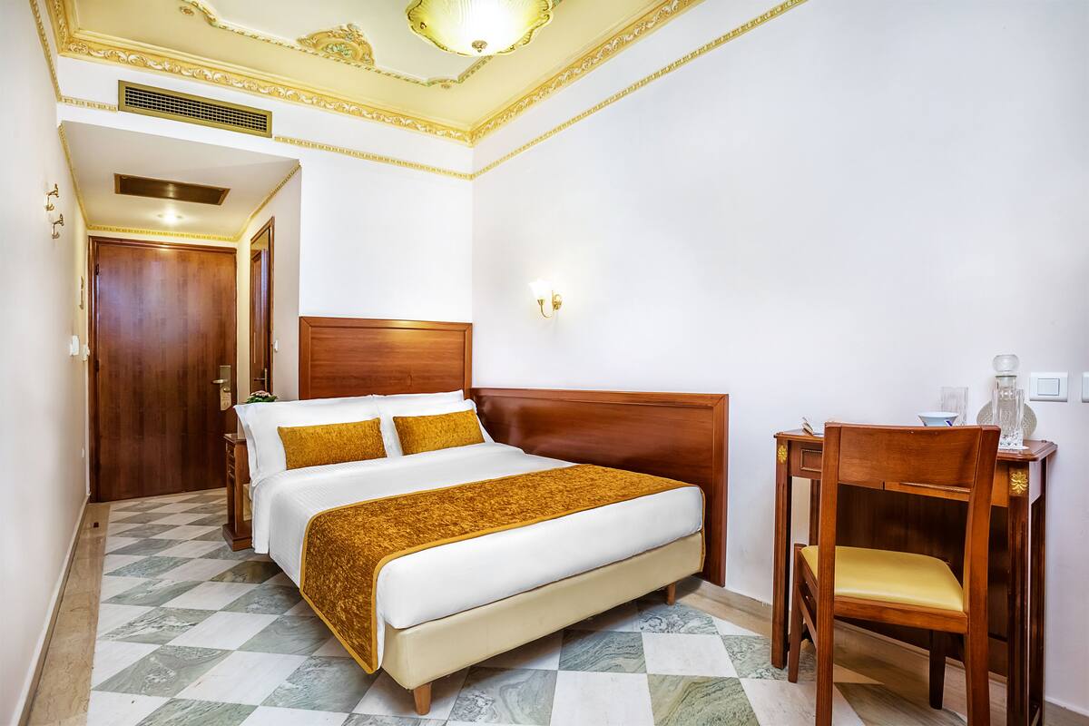 A hotel room with a large bed, wooden headboard, a wooden chair, and a small table with a lamp.