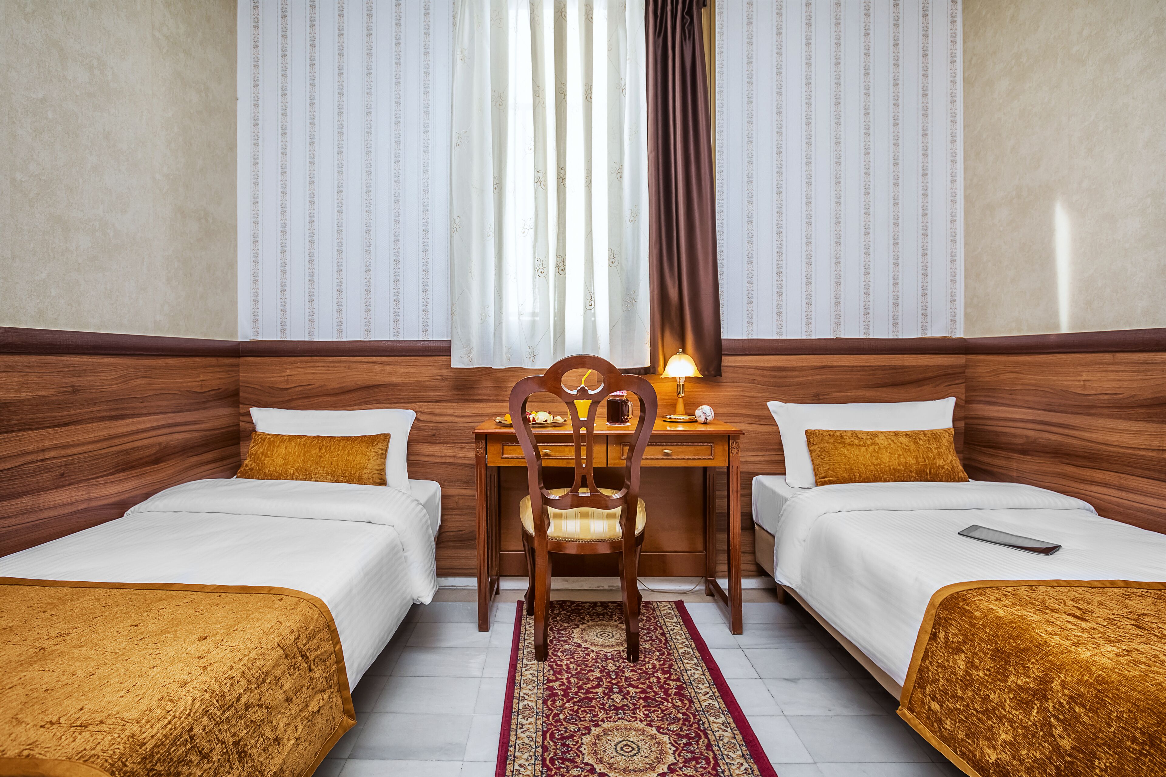 A hotel room with two single beds, a wooden desk with a chair, a lamp, and a patterned rug.