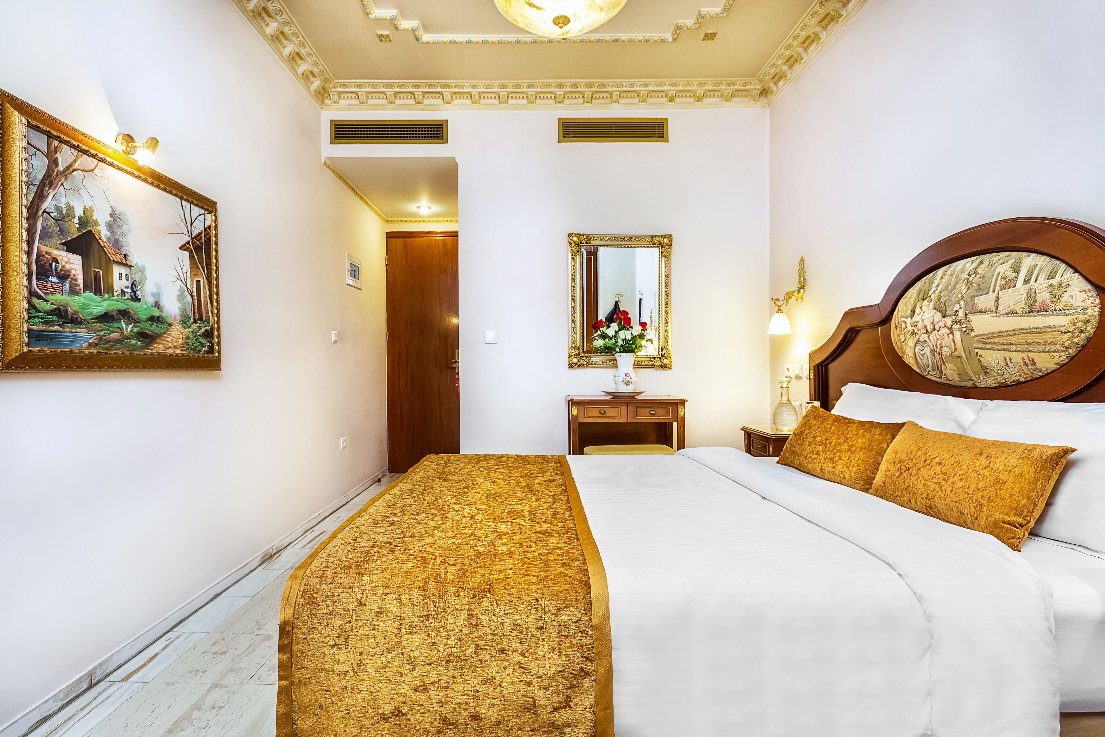 A hotel room with a large bed, a wooden headboard, a nightstand with a lamp, a mirror, and a painting on the wall.