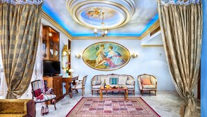 A luxurious living room with a large mural, elegant furniture, and a chandelier. - Imperial Palace Classical Hotel Thessaloniki (Thessaloniki)