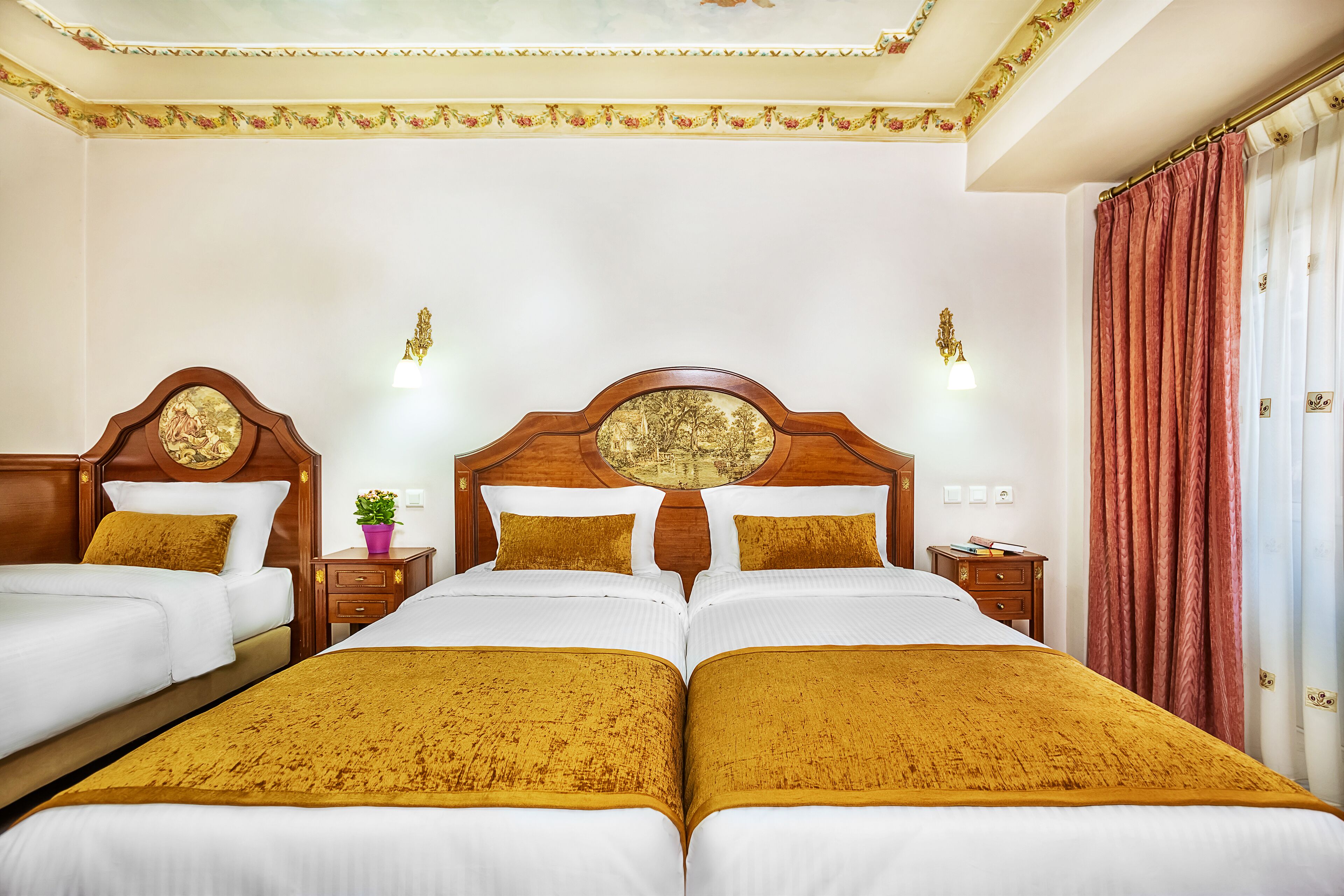 A hotel room with two beds, a wooden headboard with a circular design, a nightstand with a lamp, and a red curtain.