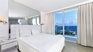 A modern hotel room with a large bed, a bedside table with a lamp, a mirror, and a view of the sea through floor-to-ceiling windows.