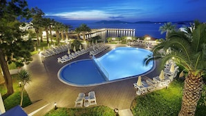 A resort pool area with lounge chairs and umbrellas, surrounded by palm trees and a view of the sea.