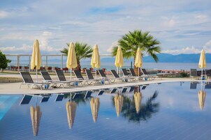 Zona junto a la piscina con tumbonas y sombrillas, vista despejada al mar y piscina tranquila que refleja el entorno.