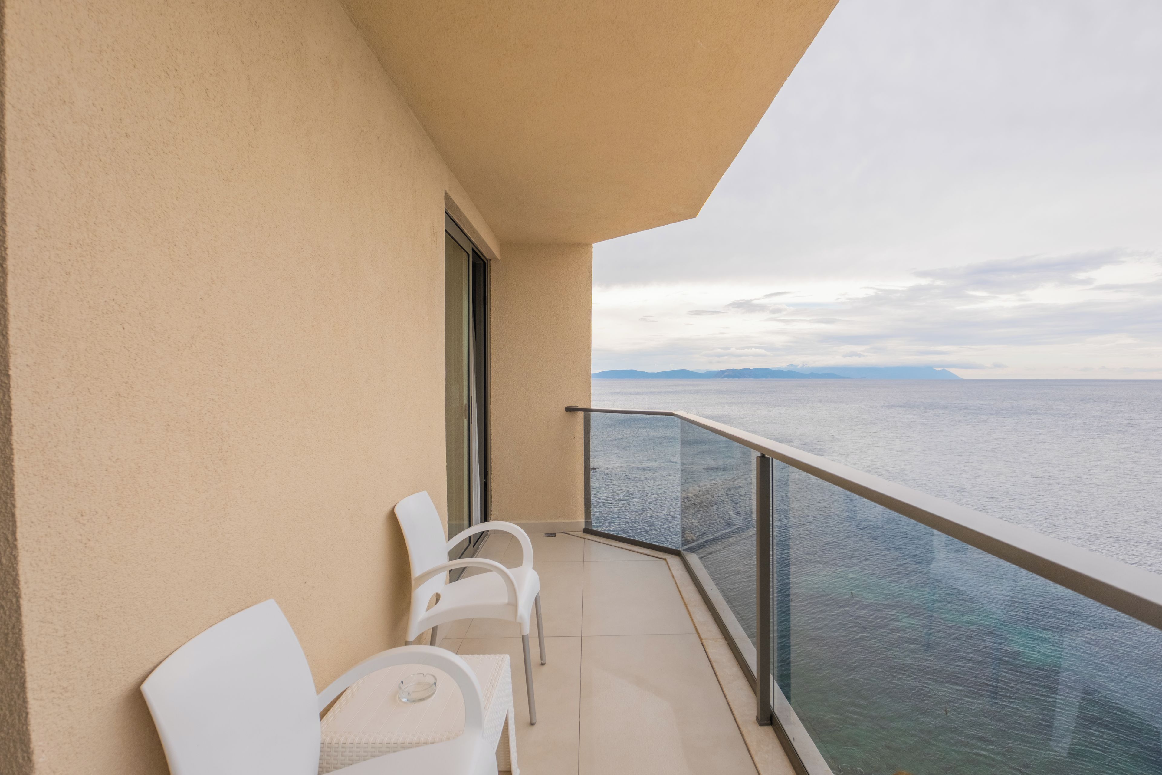 A balcony with a view of the sea, featuring two white chairs and a glass balustrade.