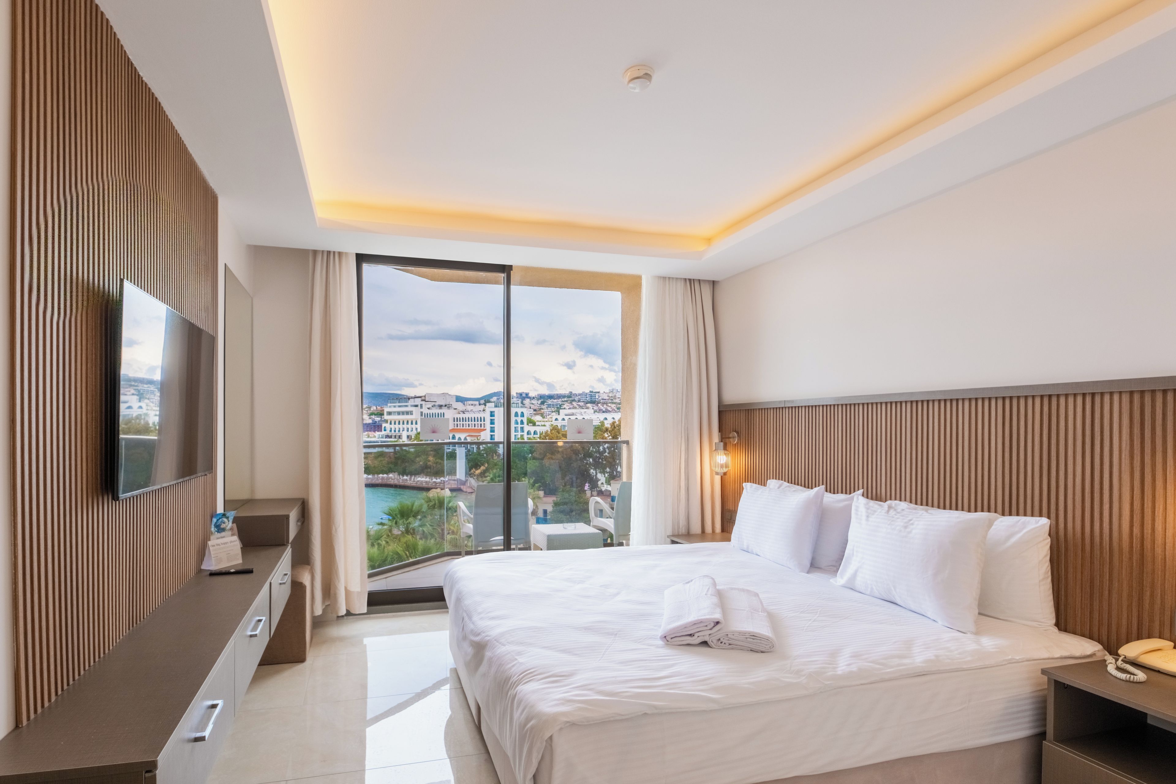 A hotel room with a large bed, a flat-screen TV, a desk with a telephone, and a view of a cityscape through a balcony.