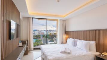 A hotel room with a large bed, a flat-screen TV, a desk with a telephone, and a view of a cityscape through a balcony.