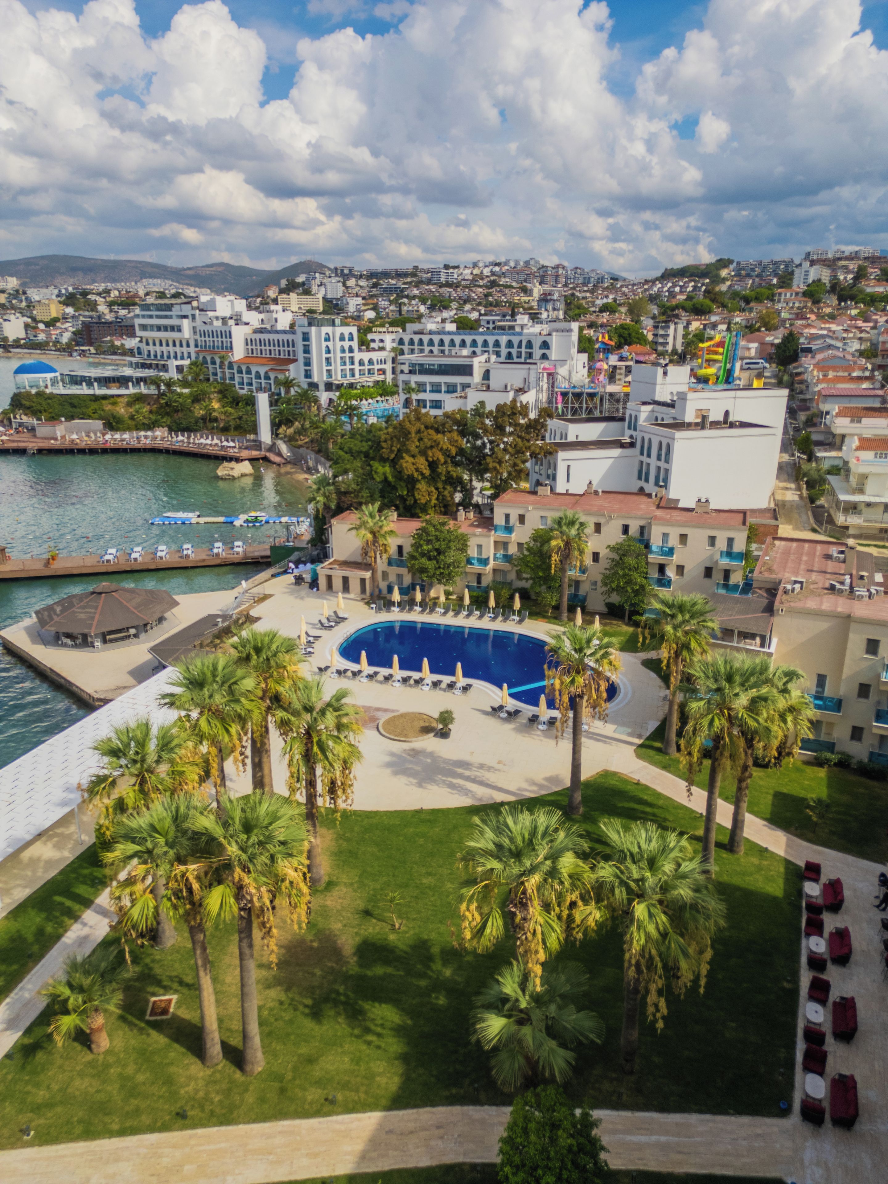 A coastal cityscape with a prominent swimming pool surrounded by palm trees and lounge chairs.