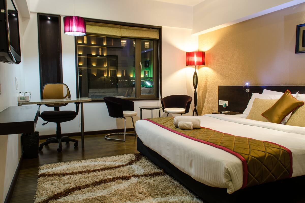 A hotel room with a bed, desk, chairs, and a window with a view of the city at night.