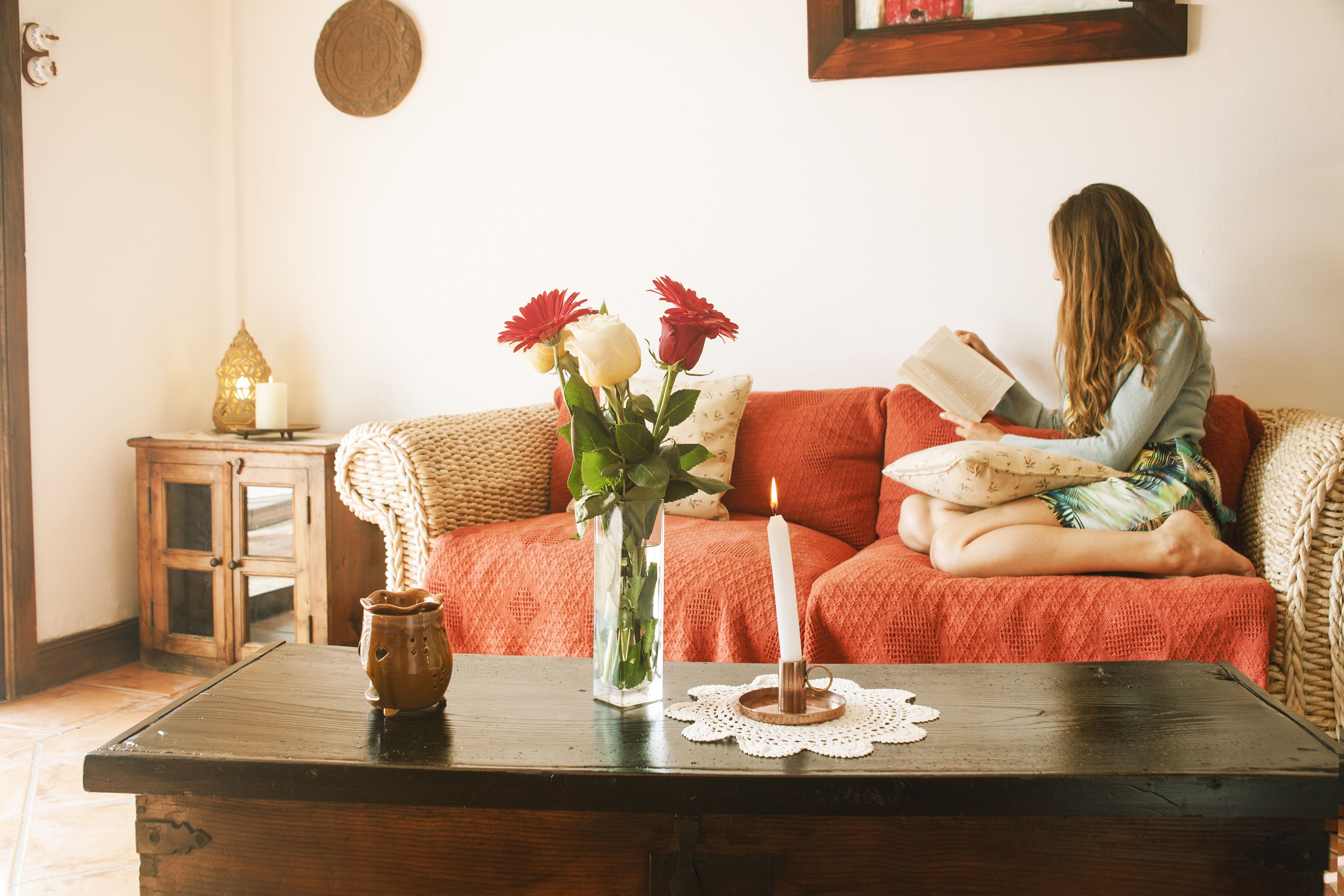 Een gezellige woonkamer met een houten bijzettafeltje, een vaas met bloemen, een brandende kaars en een persoon die een boek leest.