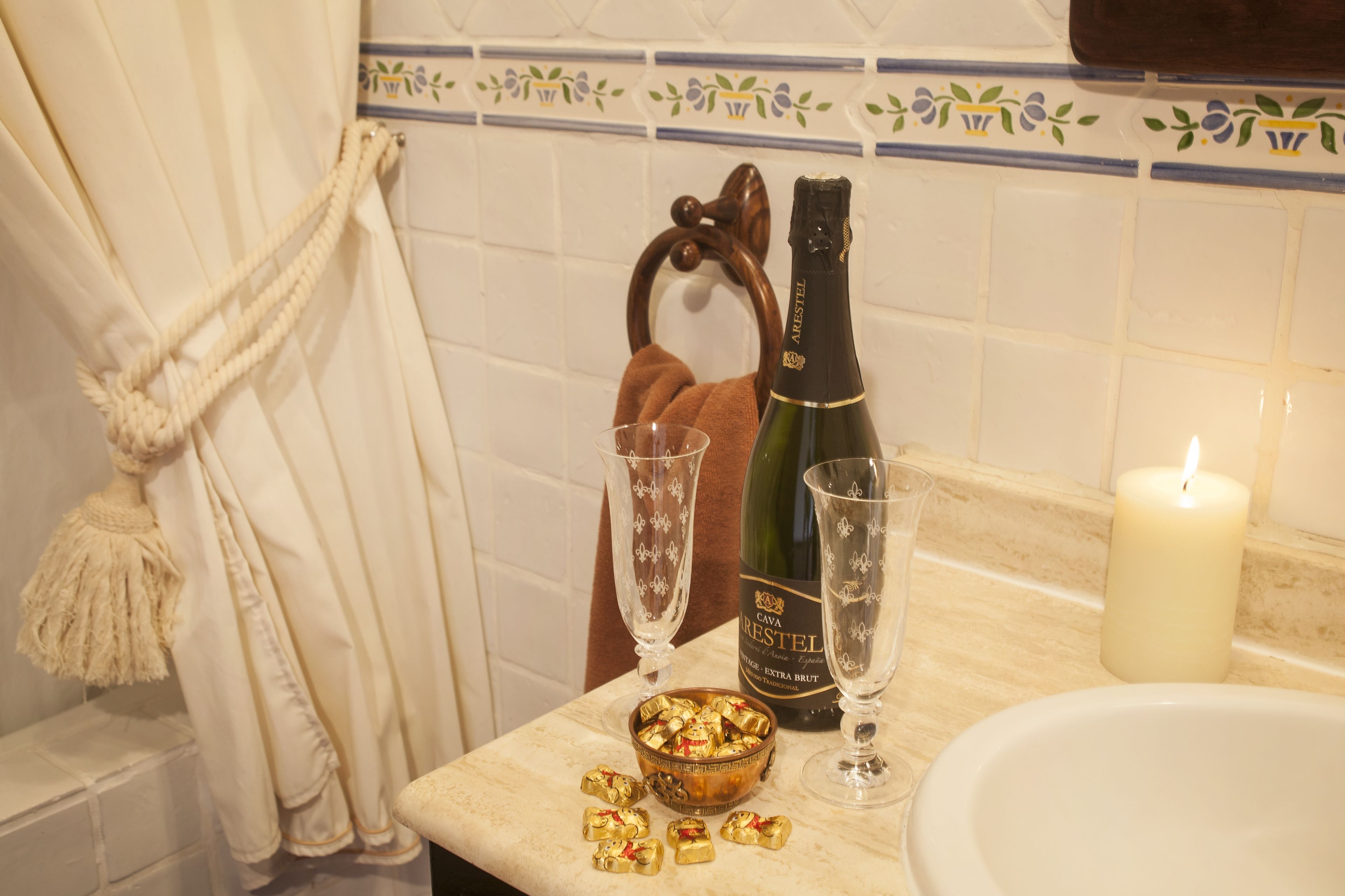 Une salle de bain avec une baignoire, un porte-serviettes, une bouteille de champagne Cava Restel, deux verres et un bol de chocolats.