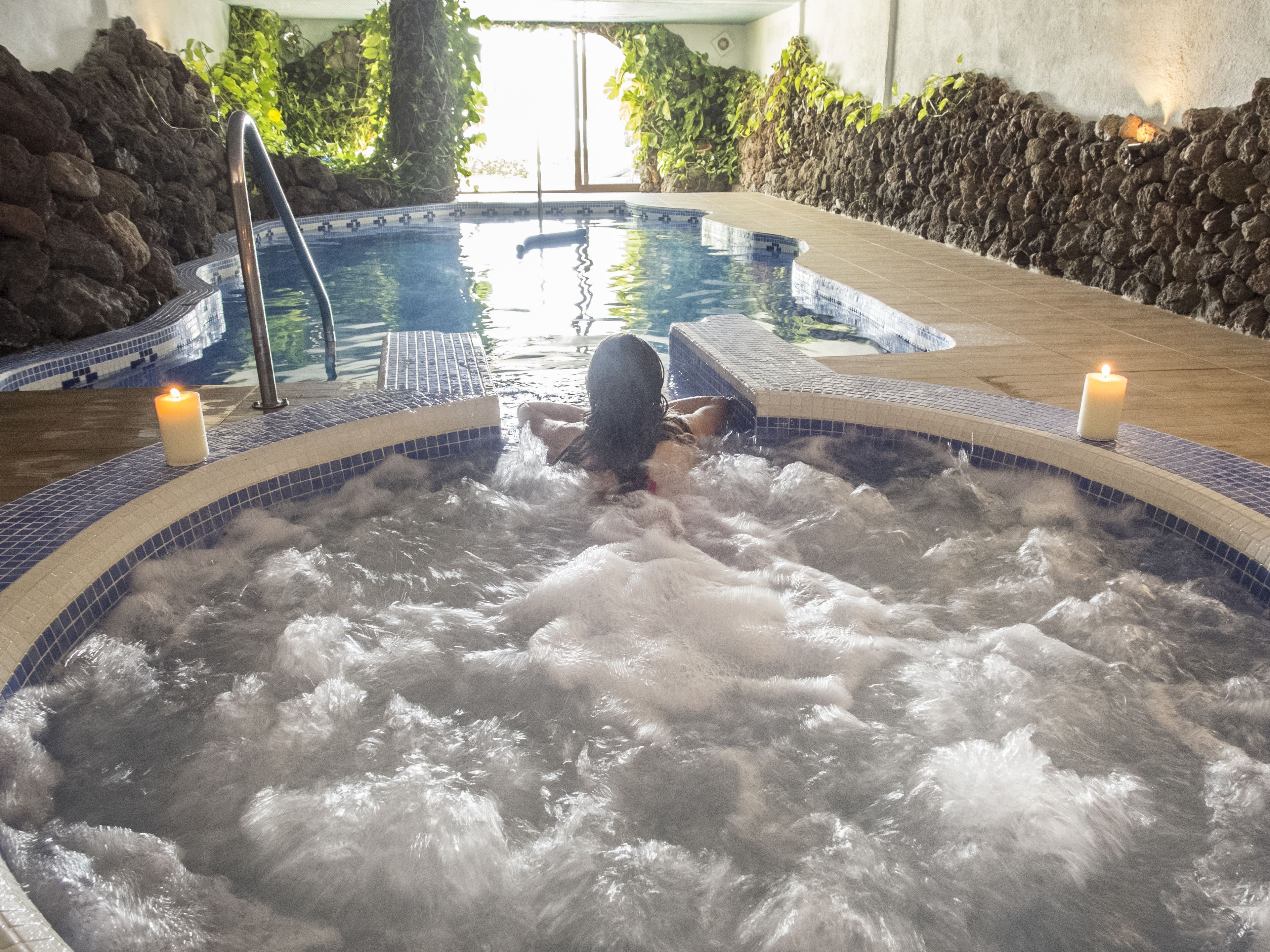 Ein Whirlpool mit zwei brennenden Kerzen, umgeben von einem gefliesten Poolbereich mit einer Steinwand und Pflanzen.