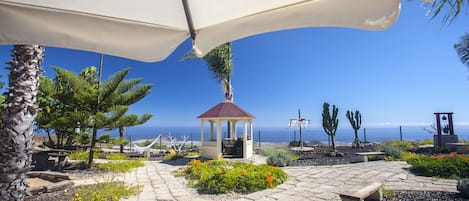A gazebo with a red roof, surrounded by a garden with various plants and flowers, and a view of the sea in the background.