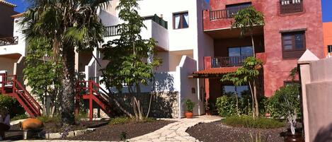 Een woonwijk met een wit gebouw, een rood gebouw en een balkon met een rode balustrade. Voor op de foto staat een palmboom en een plant in een pot.