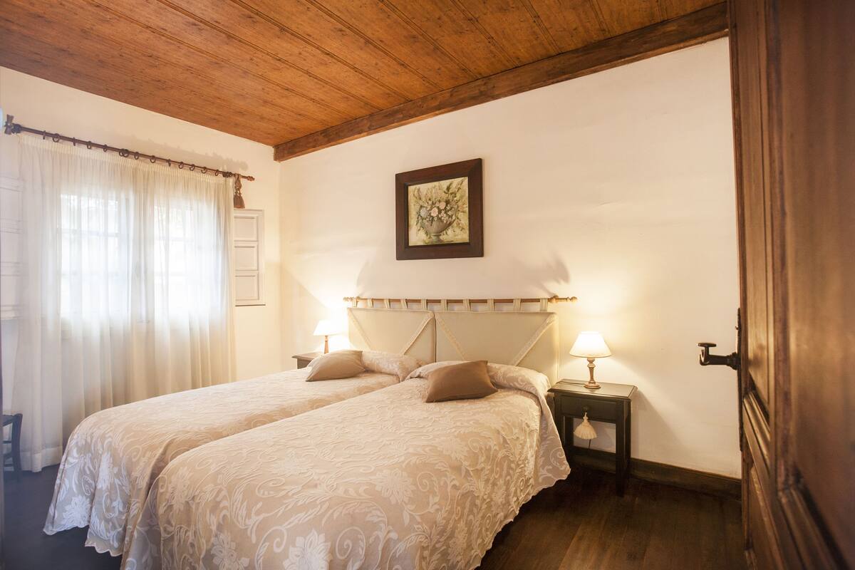 A double room with two beds, a wooden ceiling, a framed picture on the wall, and a nightstand with a lamp.